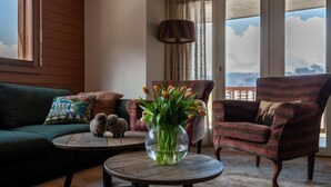 A cozy living room with a green sofa, a plaid armchair, a round wooden coffee table with a vase of tulips, and a view of mountains through large windows. - Les Chouettes 23 by Interhome (Nendaz)