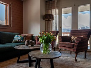 Ein gemütliches Wohnzimmer mit einem grünen Sofa, einem karierten Sessel, einem runden Holztisch mit einer Tulpenvase und Blick auf Berge durch große Fenster.