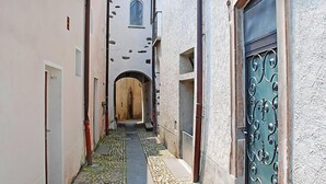 Ein schmaler Gang mit Kopfsteinpflasterboden, einer Holztür am Ende und einem Metallgitter auf der rechten Seite.