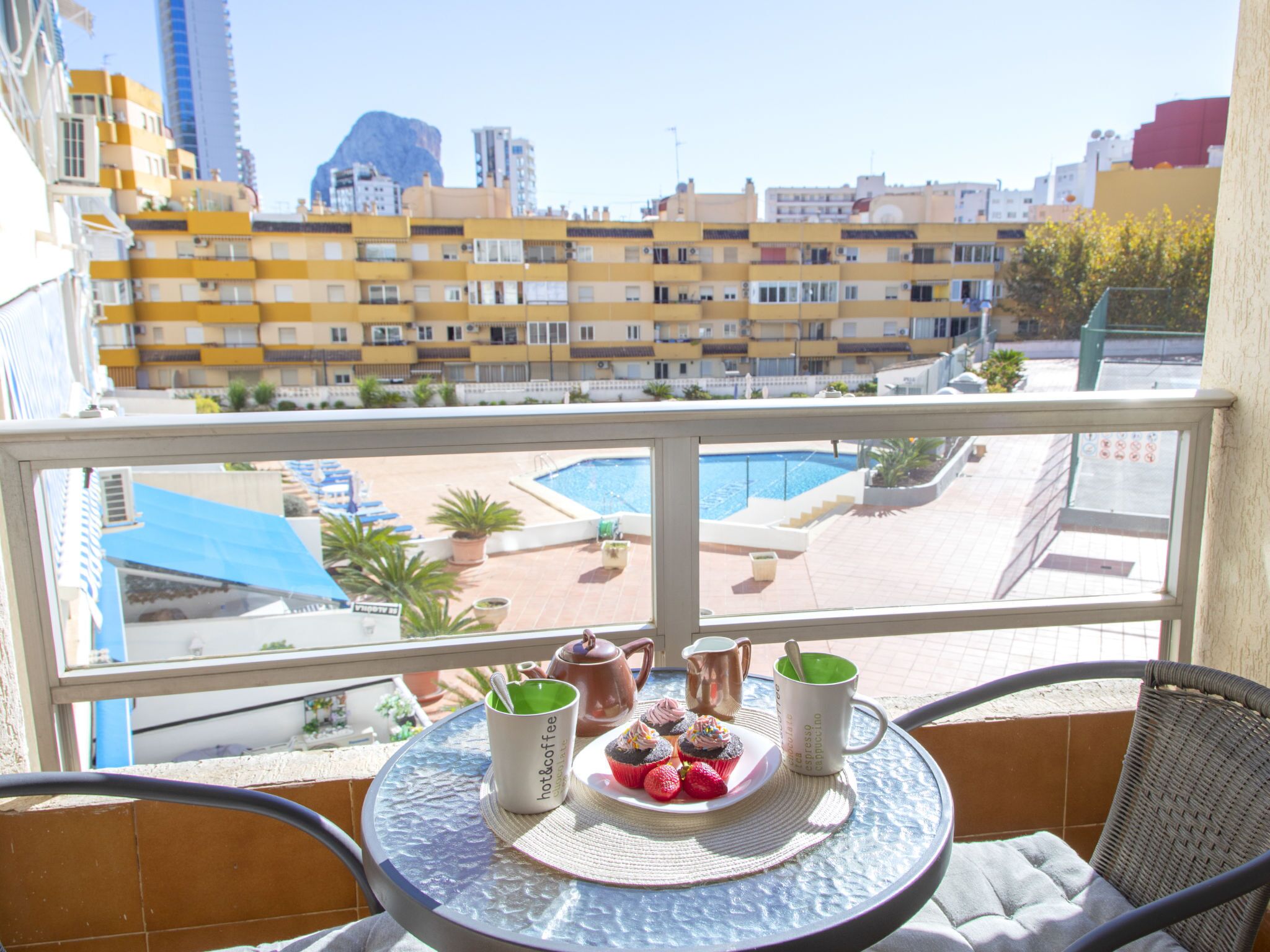A balcony with a table set for breakfast, overlooking a swimming pool and a cityscape.