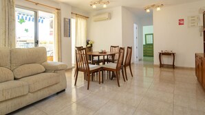 Un salon comprenant un canapé, une table à manger et des chaises. On y trouve une fenêtre avec des rideaux, une porte et une petite table sur laquelle est posée une plante.