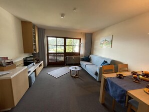 A compact living space with a kitchenette, a sofa, a dining table, and a large window offering a view of greenery.