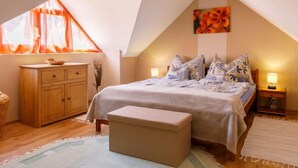 Un dormitorio con techo inclinado, una cama con almohadas estampadas, un aparador de madera y una ventana con cortinas translúcidas.