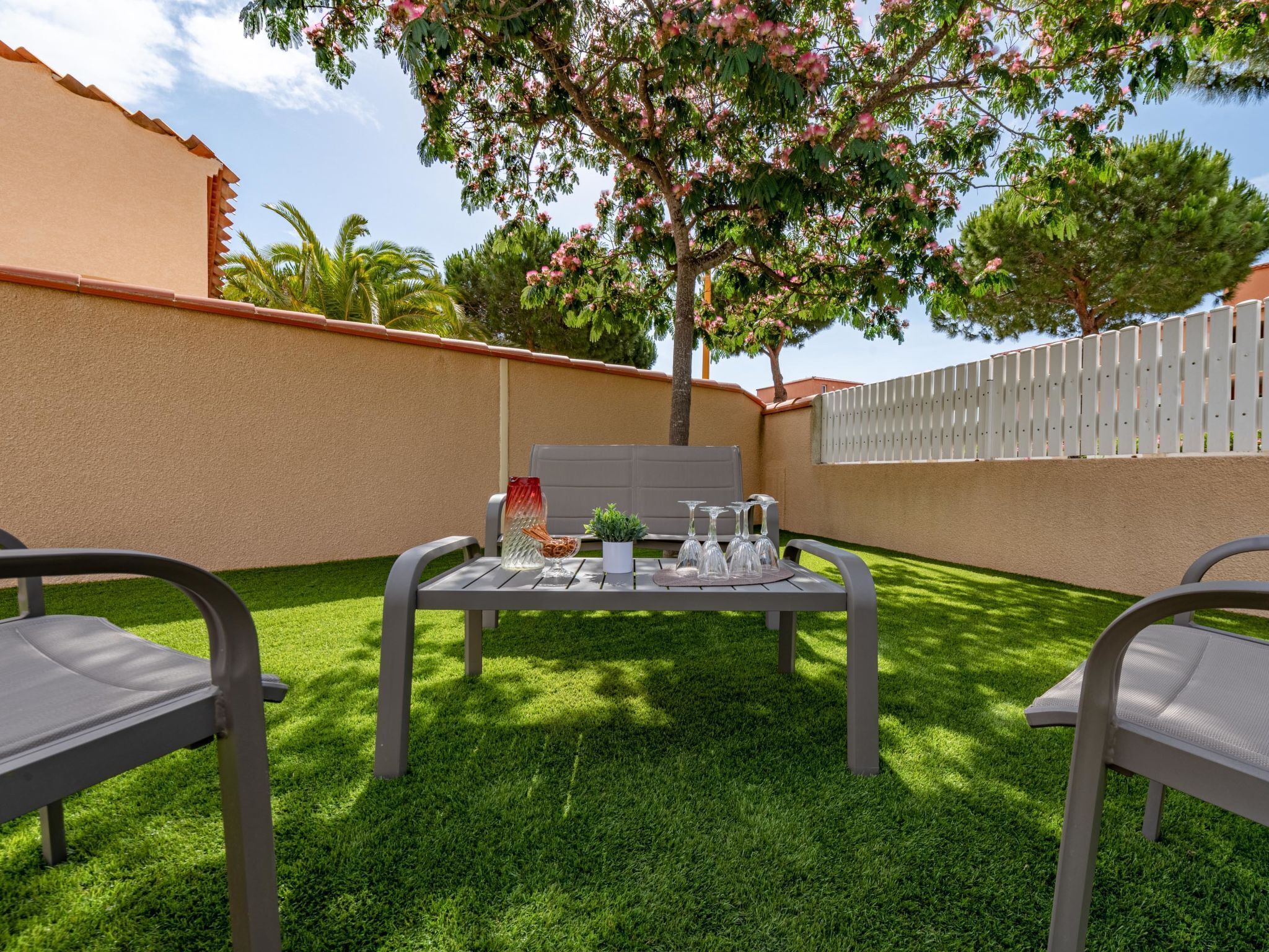 A patio with a table set for dining, surrounded by chairs, and a white fence in the background.