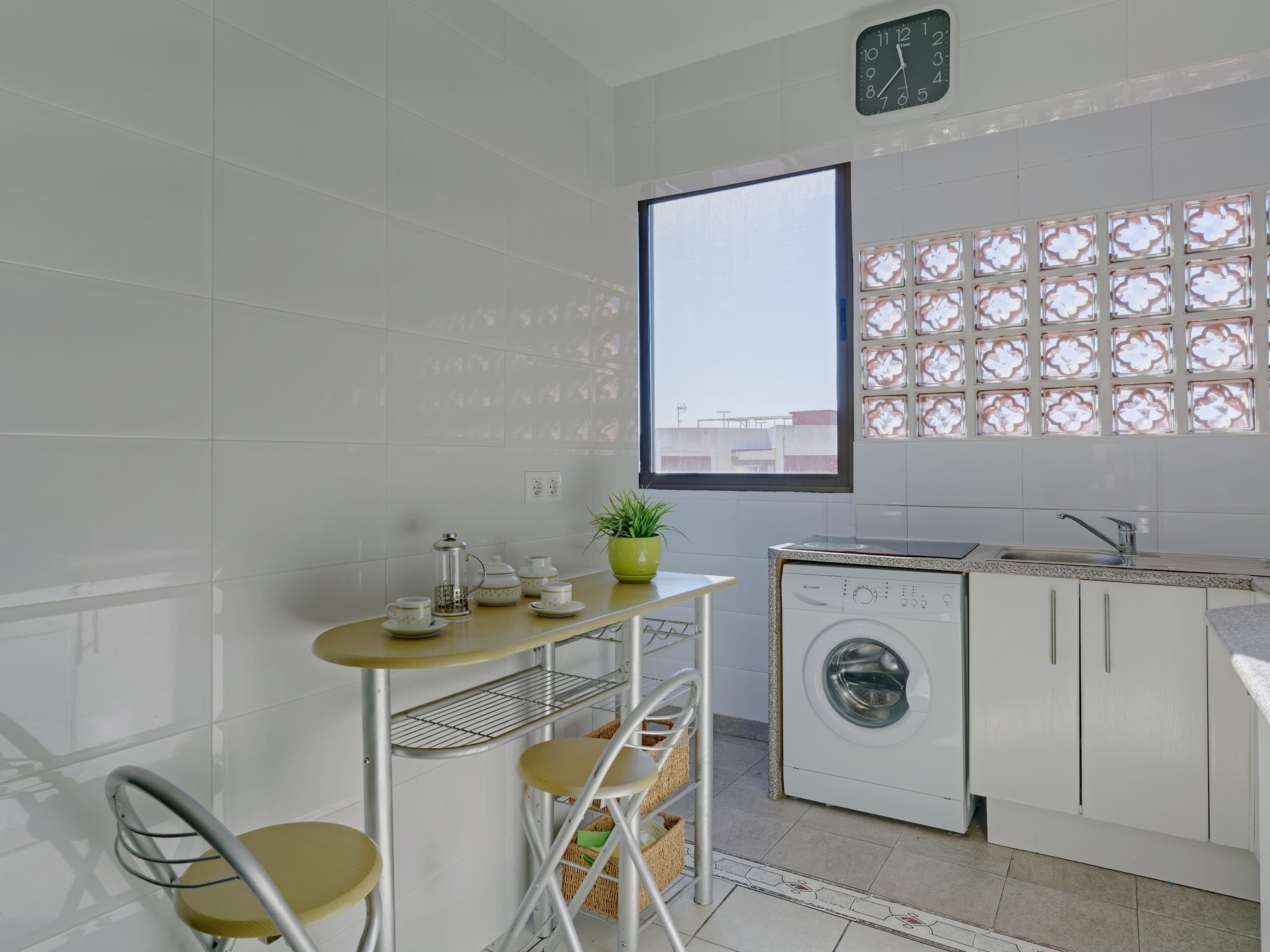 Uma cozinha compacta com máquina de lavar, uma mesa pequena com cadeiras e uma janela com painéis de vidro decorativos.