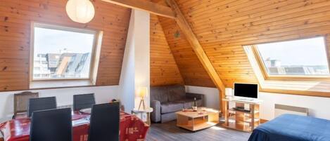 Een knus zolderkamertje met een eettafel, stoelen, een bank, een televisie en een groot raam.