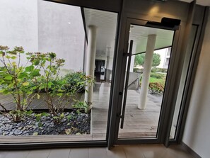 A modern building with a glass door leading to a covered outdoor area with plants and a stone pathway.
