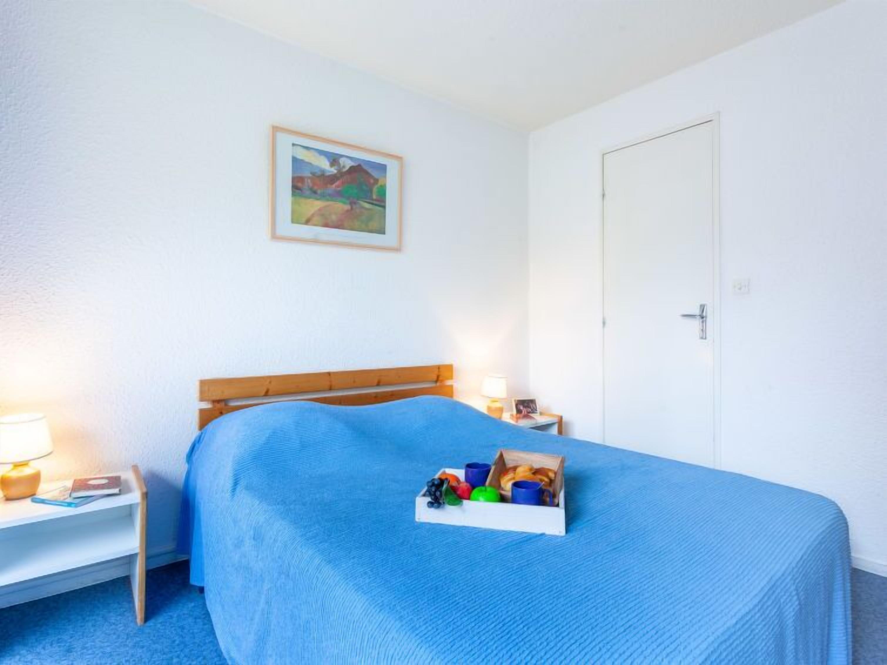 Une chambre avec un lit simple, une table de chevet, une lampe, un tableau encadré au mur et une porte.