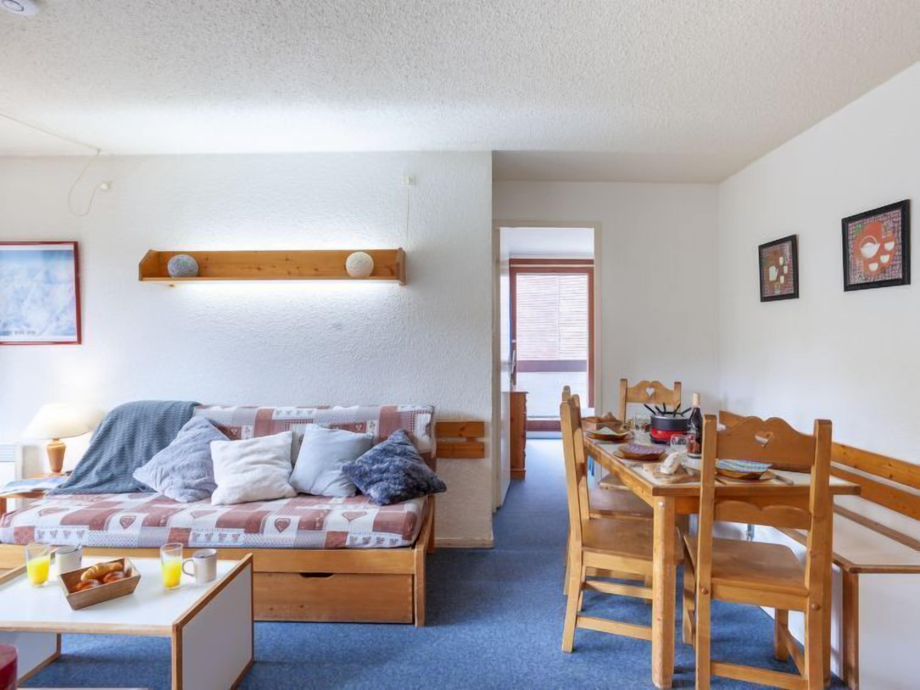 Een knus interieur met een bed, een eettafel en stoelen. Tegen de muur staat een plank met decoratieve objecten.