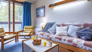 Una habitación acogedora con un sofá de madera, dos sillones, una pequeña mesa con productos para el desayuno y un ventanal con cortinas.