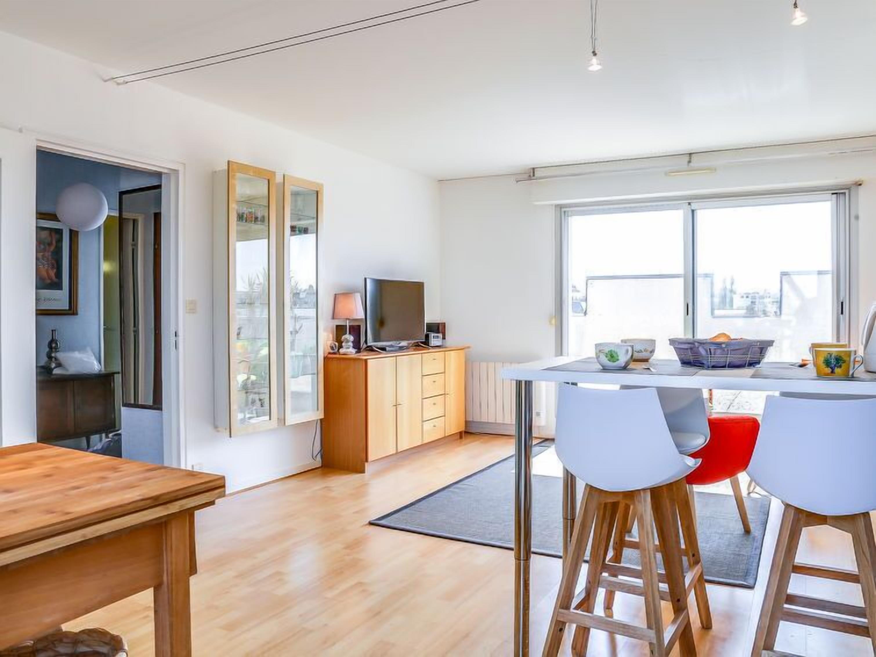A modern kitchen with a dining area, wooden cabinets, a TV, and large windows.