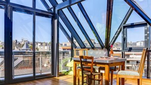 Uma área de refeições com mesa e cadeiras de madeira, janelas amplas e vista para edifícios.