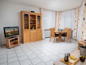 A living room with a TV, wooden cabinet, dining table with chairs, and a small kitchen area with a bottle of champagne.