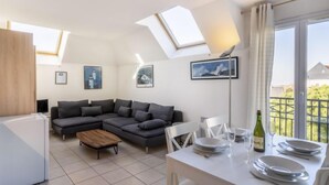 Ein modernes Wohnzimmer mit einem grauen Ecksofa, einem Holztisch und einem Essbereich mit einem weißen Tisch, gedeckt für zwei Personen.