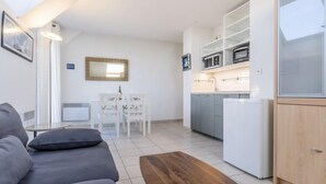 Ein modernes Wohnzimmer mit einem grauen Sofa, einem Holztisch und einem Essbereich mit weißem Tisch und Stühlen. Im Hintergrund ist eine Küche mit weißen Schränken und einer Mikrowelle zu sehen.