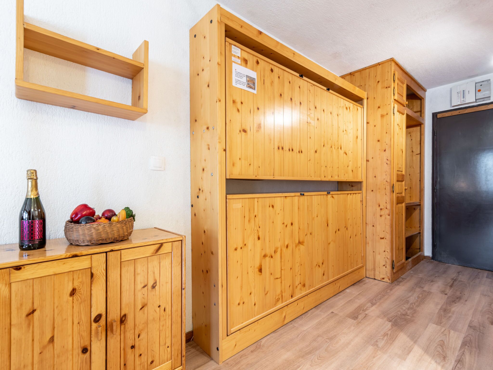 A wooden cabinet with a bottle and fruit basket, a wooden shelf, and a black refrigerator.