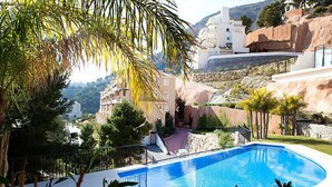En swimmingpool med en buet kant, omgivet af grøn vegetation og et bjergrigt landskab.