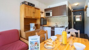 A compact kitchen with wooden cabinets, a microwave, and a stove. There's a dining table with a brochure, a bottle of juice, and a bowl. - Vanoise 152 by Interhome (Val Thorens)