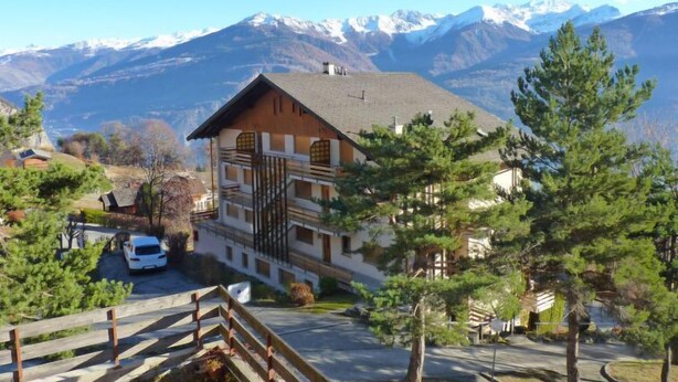 A mountain lodge with a wooden facade, a staircase leading to the entrance, and a view of snow-capped mountains in the background. - Centaure A RS 3 by Interhome (Ovronnaz)