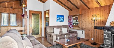 A cozy living room with a wooden ceiling, a fireplace, a sofa, and a dining table with wine glasses.