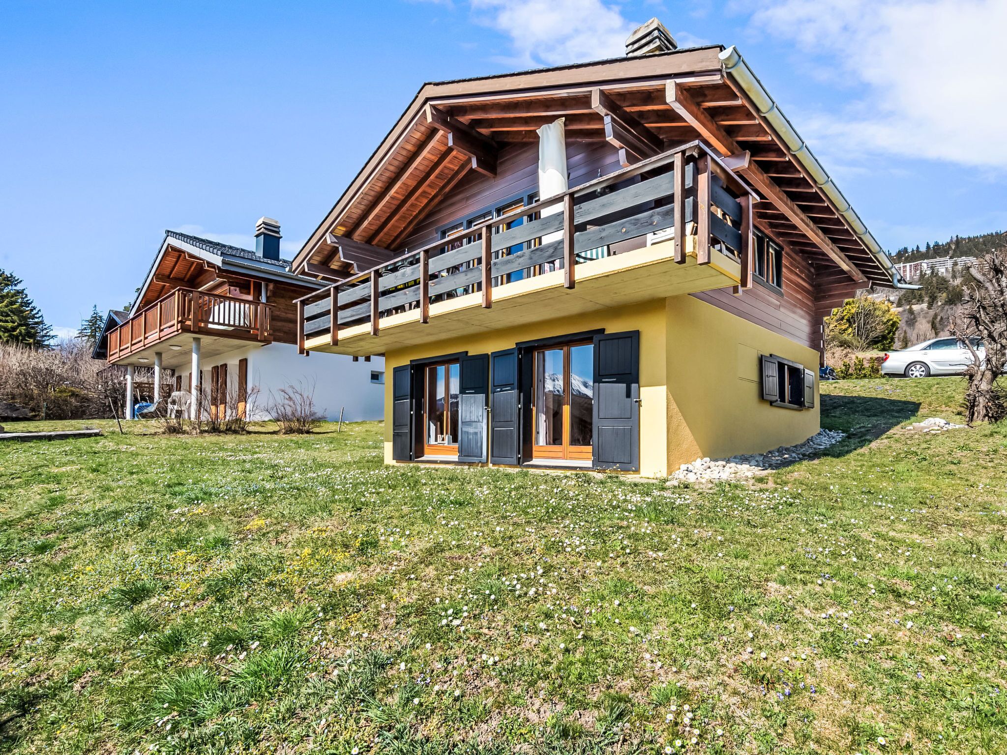 A two-story chalet with a balcony, large windows, and a grassy yard.