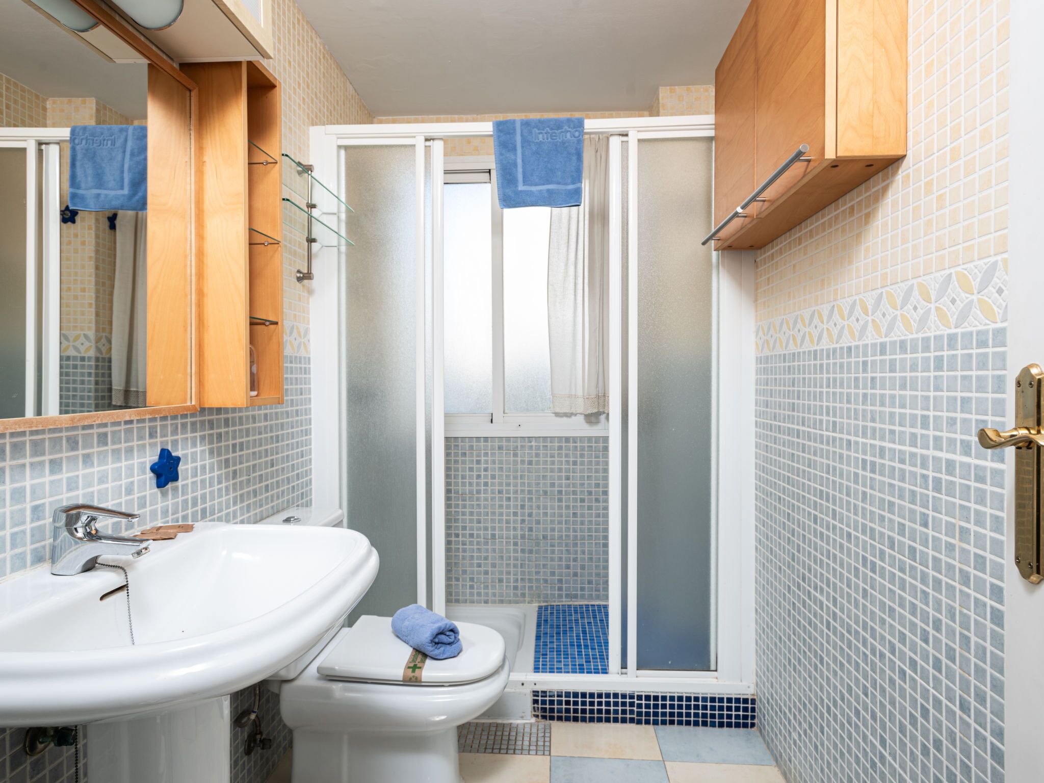A bathroom with a white sink, toilet, and shower with a glass door. There are blue towels hanging on the door handles.