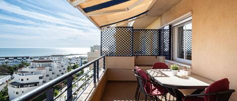 A balcony with a table and chairs, a view of the sea, and a cityscape in the background.