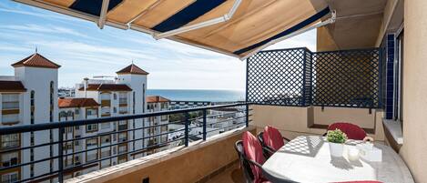 Uma varanda com mesa e cadeiras, vista para edifícios e para o mar, e uma área coberta por toldo.