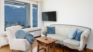 Un salon chaleureux avec un canapé et un fauteuil, un téléviseur fixé au mur et une vue sur les montagnes par une grande fenêtre.