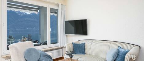 A cozy living room with a sofa and armchair, a TV mounted on the wall, and a view of mountains through a large window.