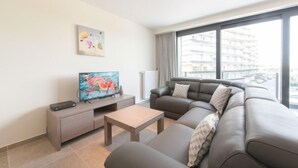 Uma sala de estar moderna com um sofá cinzento, uma mesa de centro de madeira e uma televisão de tela plana em um suporte. Há um quadro na parede e uma janela com vista para edifícios.