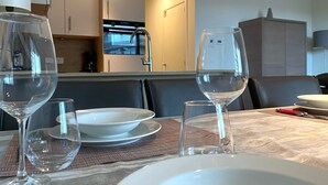 Mesa de jantar posta com taças de vinho e pratos, cozinha ao fundo e um armário.