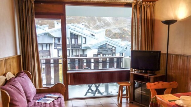 Ett rum med en soffa i trä, ett litet bord med en duk på, en tv på ett stativ och en balkong med utsikt över byggnader.