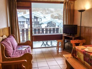 Une pièce avec un canapé en bois, une petite table recouverte d’une nappe, un téléviseur posé sur un meuble et un balcon offrant une vue sur les bâtiments.