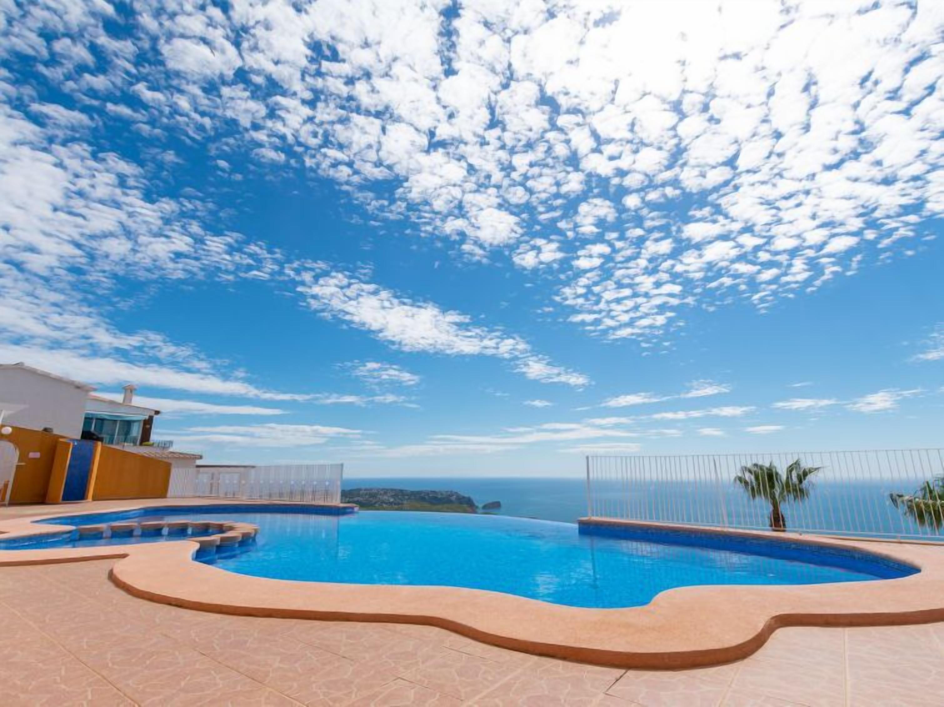 A swimming pool with a curved edge, a clear blue sky, and a distant view of the sea.