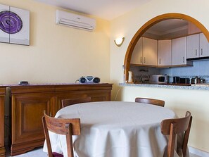 A dining area with a round table, wooden chairs, a kitchen with cabinets and a microwave, and an air conditioning unit on the wall.