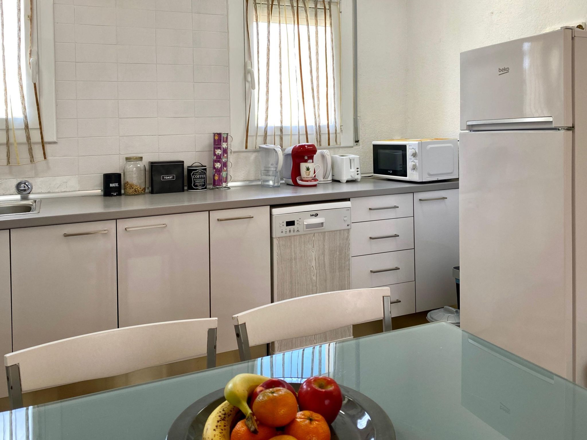Una cocina moderna con armarios blancos, lavavajillas, microondas y tostadora. Hay una mesa de vidrio con un frutero que contiene manzanas, naranjas y un plátano.