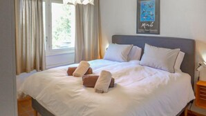 Una cama bien hecha con ropa blanca y toallas enrolladas, un cabecero gris y una obra enmarcada en la pared.