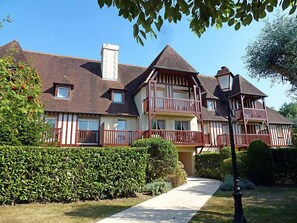 A large, multi-story house with a prominent balcony on the second floor, surrounded by well-maintained hedges and a clear sky.