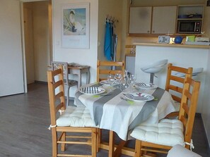 A dining area with a table set for four, wooden chairs, a painting on the wall, and a kitchen in the background.