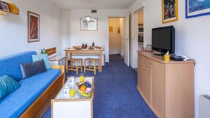Een ruimtelijk ingerichte leefruimte met een blauwe bank, een houten eettafel, een televisie op een houten kast, en een gang die naar een andere kamer leidt.
