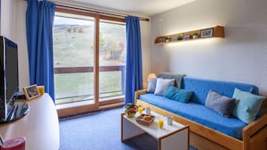 Een woonkamer met een blauwe bank, een salontafel met ontbijtartikelen, een televisie en uitzicht op een grasveld door grote ramen.