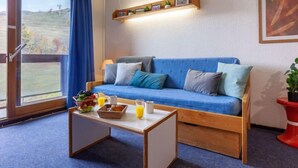Een gezellige kamer met een blauwe bank, een houten bijzettafeltje met ontbijtspullen en uitzicht door een groot raam met blauwe gordijnen.