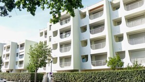 Un immeuble résidentiel moderne à plusieurs étages, doté de balcons, d’un espace de stationnement et d’une haie.