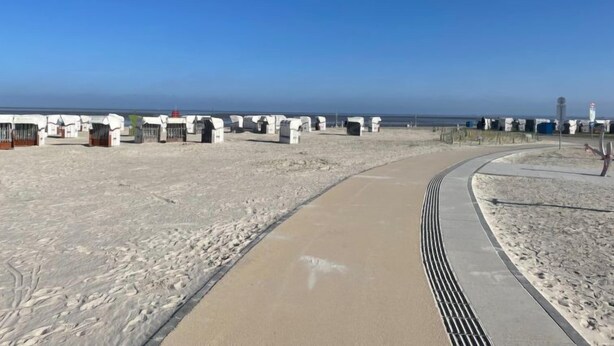 Una playa arenosa con un camino curvo que conduce a varias estructuras blancas, posiblemente cabañas de playa, a lo largo de la costa.
