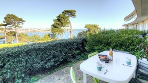 Eine Terrasse mit gedecktem Tisch für zwei Personen mit Blick auf die Küste, Bäume und ein Gewässer.
