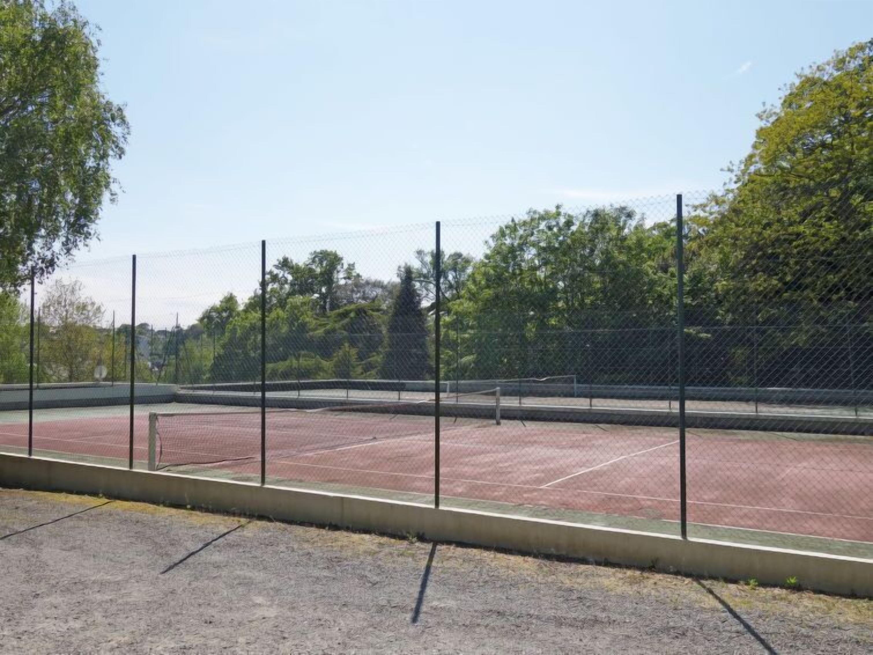 A tennis court with a chain-link fence, surrounded by trees and a clear sky.