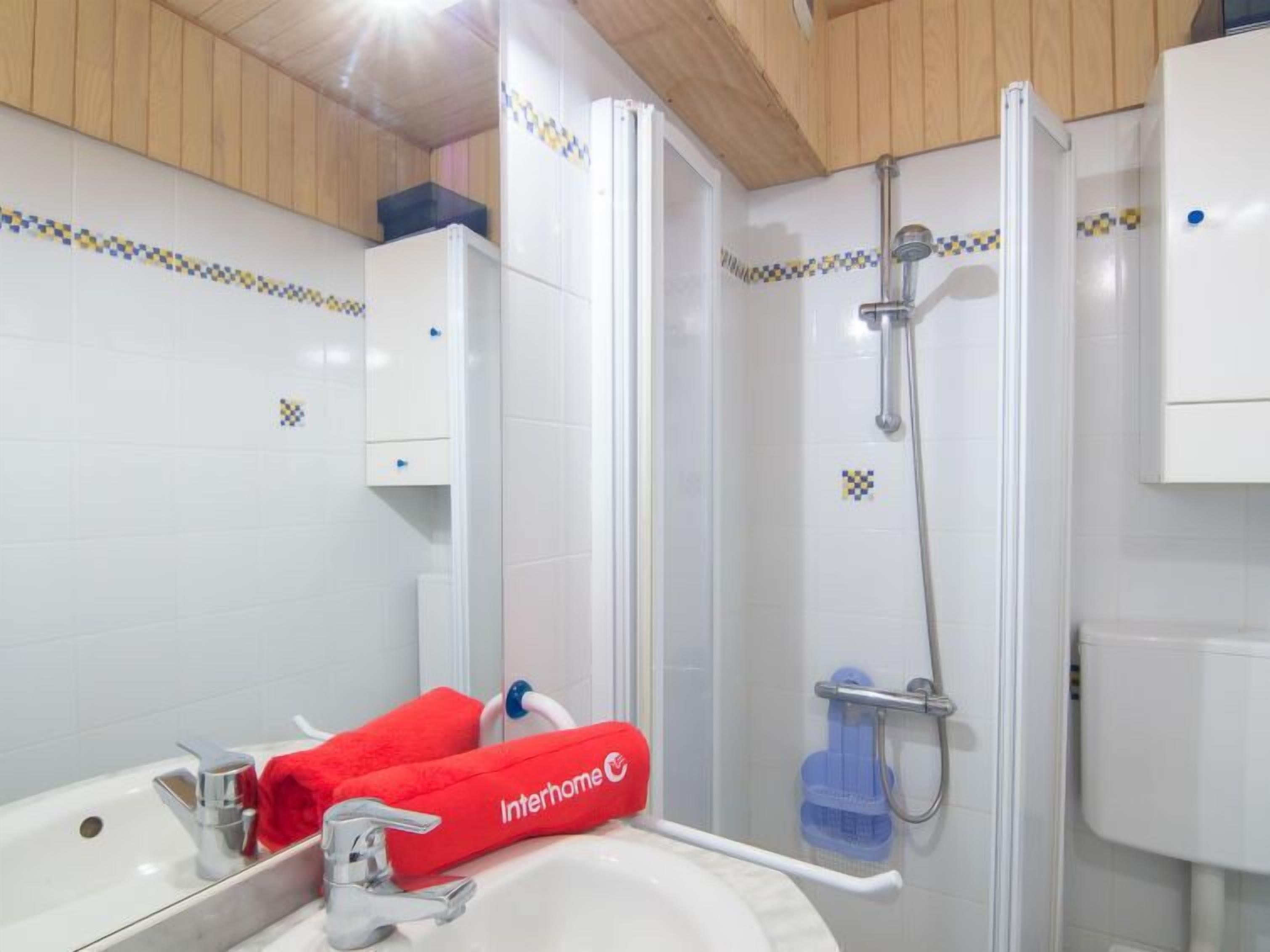 A bathroom with a sink, mirror, and shower. There are red towels and a showerhead.