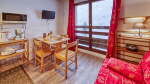 Una habitación acogedora con una mesa de comedor de madera, sillas, un sofá, un televisor instalado en la pared y una ventana con cortinas rojas.