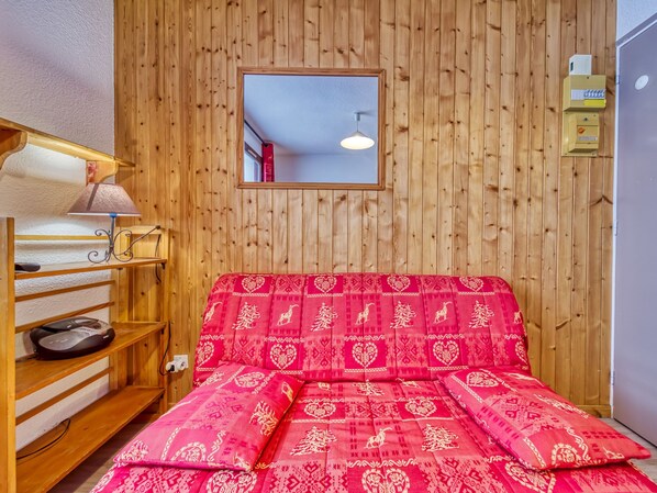 A cozy room with a wooden wall, a bed with red patterned bedding, a shelf with a lamp, and a small mirror. - Argousier 45 A by Interhome (Les Menuires)